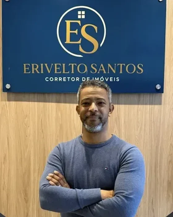 Erivelto Santos, corretor de imóveis, de braços cruzados diante da placa do escritório